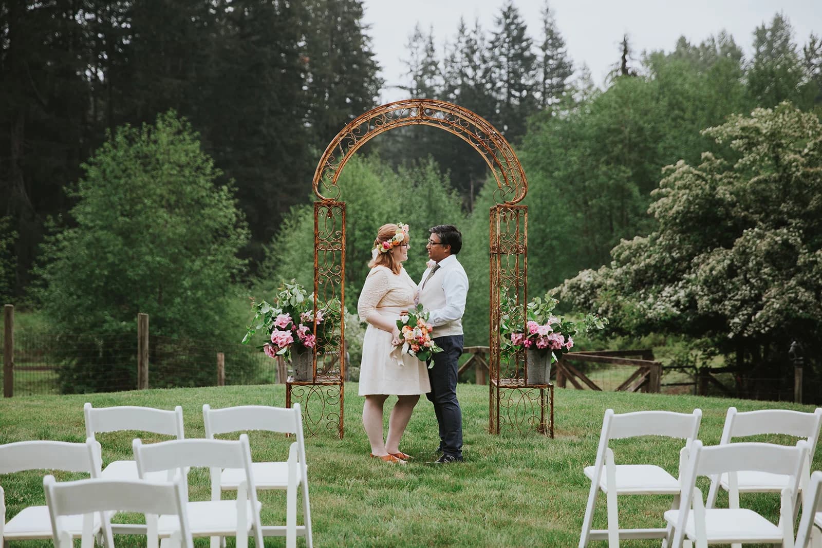 An outdoor ceremony arch dressed with flowers on open grass, white chairs set before a Pacific Northwest tree line
